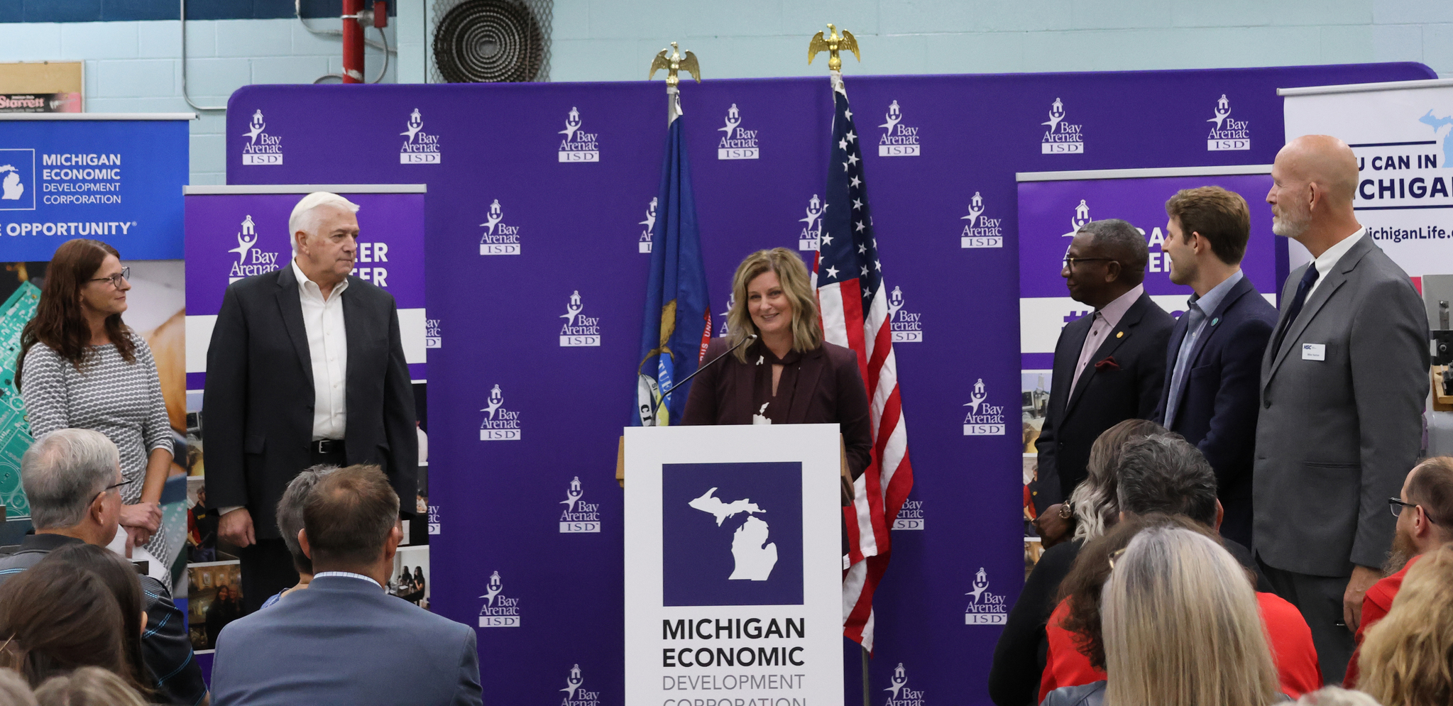 A woman speaks at a podium with a Michigan Economic Development Corporation sign, surrounded by officials and attendees during a Bay Arenac ISD event held in a workshop setting.